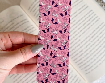 Puede incluir: Marcapáginas rosa con un patrón repetido de murciélagos blancos, lunas y libros. Los murciélagos vuelan sobre los libros y las lunas.