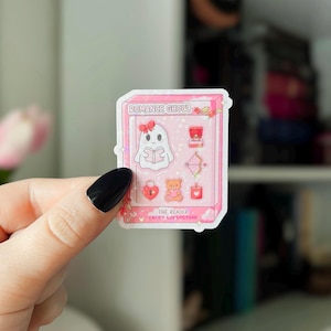 May include: A pink sticker featuring a ghost reading a book, with the text "ROMANCE GHOUL" and "THE READER TROPE COLLECTION." Other images include a ring, bow and arrow, heart lock, teddy bear, and candle.