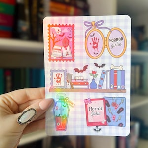 May include: A sheet of stickers with a horror theme. The stickers feature images of a handprint, a coffin-shaped bottle, a bookshelf, and the words "Horror Girlie". The stickers have a holographic effect and are on a checkered background.