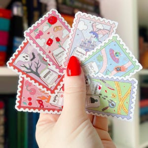 Puede incluir: Una mano sostiene una colecci&oacute;n de pegatinas coloridas con dise&ntilde;os de sellos postales. Cada pegatina presenta un tema diferente, incluyendo clubes de lectura de romance, fantas&iacute;a, terror, ciencia ficci&oacute;n, thriller y picante. Las pegatinas tienen un borde blanco y bordes decorativos.