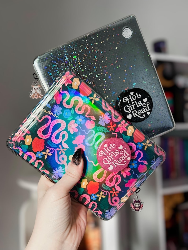 May include: Two phone cases, one clear with a glittery black design and the other with a colorful floral and snake pattern. Both cases have the text "Hot Girls Read". Each case has a charm attached.