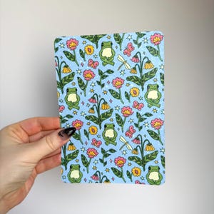 May include: A rectangular card with a light blue background featuring a repeating pattern of green frogs, pink and yellow flowers, butterflies, dragonflies, and stars. The card has rounded corners.