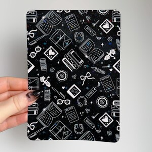 May include: A black rectangular item featuring a white pattern of books, pens, glasses, and bows. The design includes the word "BOOKS" and small stars. The item is decorated with hearts and other book-related elements.