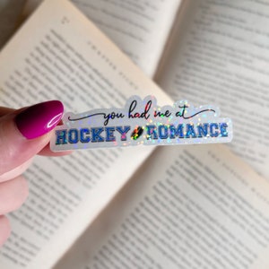 Puede incluir: Una pegatina hologr&aacute;fica con el texto "You had me at hockey romance" en escritura negra con un gr&aacute;fico de disco de hockey azul.