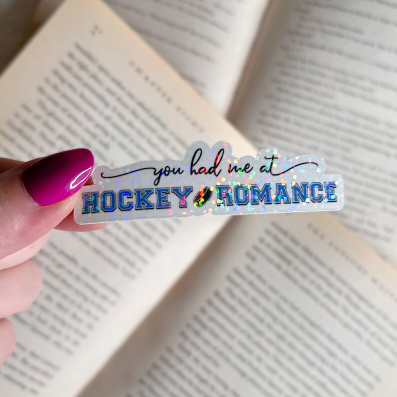 Puede incluir: Una pegatina hologr&aacute;fica con el texto "You had me at hockey romance" en escritura negra con un gr&aacute;fico de disco de hockey azul.