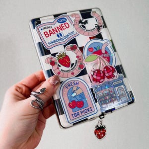 May include: A clear plastic case with a checkered pattern and various colorful stickers. The stickers feature a strawberry, cherries, a bookworm, a bookstore, and a sign that says "100% Banned Forbidden Content".