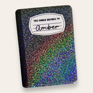 Puede incluir: Un cuaderno negro y plateado con purpurina con una etiqueta blanca que dice "This Kindle Belongs To Amber".