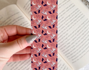 Puede incluir: Un marcapáginas rosa con un patrón repetido de murciélagos blancos, lunas y rosas. Los murciélagos sostienen libros abiertos con un unicornio blanco en cada libro.