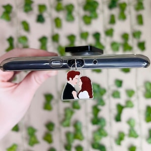 May include: A clear phone case with a black phone ring holder and a charm featuring a cartoon couple kissing. The charm is attached to the phone case with a silver ring.