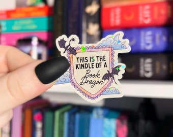 May include: A holographic sticker with a pink and blue background and a white shield with the text "This is the Kindle of a Book Dragon". The sticker features a black silhouette of a dragon with wings.
