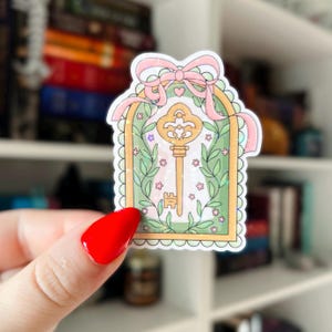 May include: A sticker featuring a gold key inside a green arched frame, adorned with pink ribbon and floral accents. The key has intricate details, and the frame is outlined with a scalloped edge. The sticker is held by a hand with red nail polish.