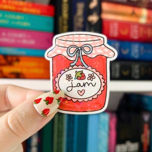 May include: A sticker of a red and pink jar of jam with a white label that says "jam" and a small strawberry on it. The jar has a blue and white gingham lid with a blue bow.