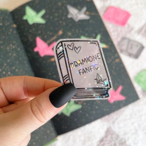 May include: A clear sticker with a book design and glitter. The book has a black outline and the text "DRAMIIONE FANFIC" is written on the cover.