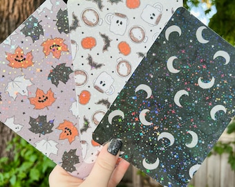 May include: Three clear, iridescent, rectangular stickers with Halloween-themed designs. The first sticker has a pattern of orange and brown leaves with jack-o-lantern faces. The second sticker has a pattern of white ghosts and coffee cups with bats. The third sticker has a black background with white crescent moons and stars.