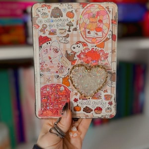 May include: A rectangular tablet case featuring a collage of autumn-themed stickers, including Snoopy, pumpkins, books, and the text "all booked for fall" and "fall girly". A heart-shaped shaker charm filled with iridescent glitter is attached.