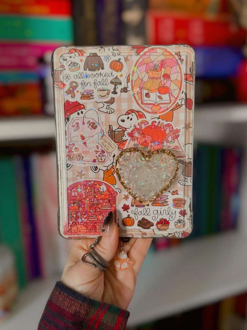 May include: A rectangular tablet case featuring a collage of autumn-themed stickers, including Snoopy, pumpkins, books, and the text "all booked for fall" and "fall girly". A heart-shaped shaker charm filled with iridescent glitter is attached.