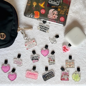 May include: A collection of 13 colorful bookish charms with various designs, including a pink heart with the text "Rhysand", a pink heart with the text "Umbra Mortis", a pink ticket with the text "Admit One", a black ticket with the text "Admit One Dark Academia", a pink heart with the text "Xaden Riorson", a pink heart with the text "Beautiful Disaster", a pink ticket with the text "Never Not Reading Smut", a pink ticket with the text "Situational", a pink ticket with the text "True Crime Girlie", a black ticket with the text "Romanstasy Book Club", a white origami crane, a green frog, and a pink ticket with the text "The Midnight Book Club".