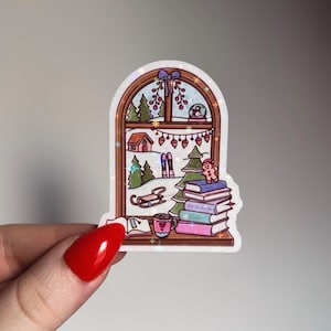 May include: A sticker featuring a winter scene viewed through a window. The scene includes a snow globe, trees, a house, skis, a sled, and a stack of books. A gingerbread man and a mug of hot chocolate are also present.