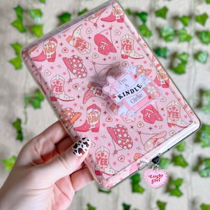May include: A pink and white clear case with a glittery design of cowboy boots, hats, and stars. The case has a pink charm that says "Lucky Girl" and a pink sticker that says "I'm a Kindle Girlie".