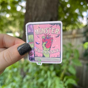 May include: A pink and green sticker with a book cover design. The book cover says "Monster Romance" and features a green hand holding a bloody heart.