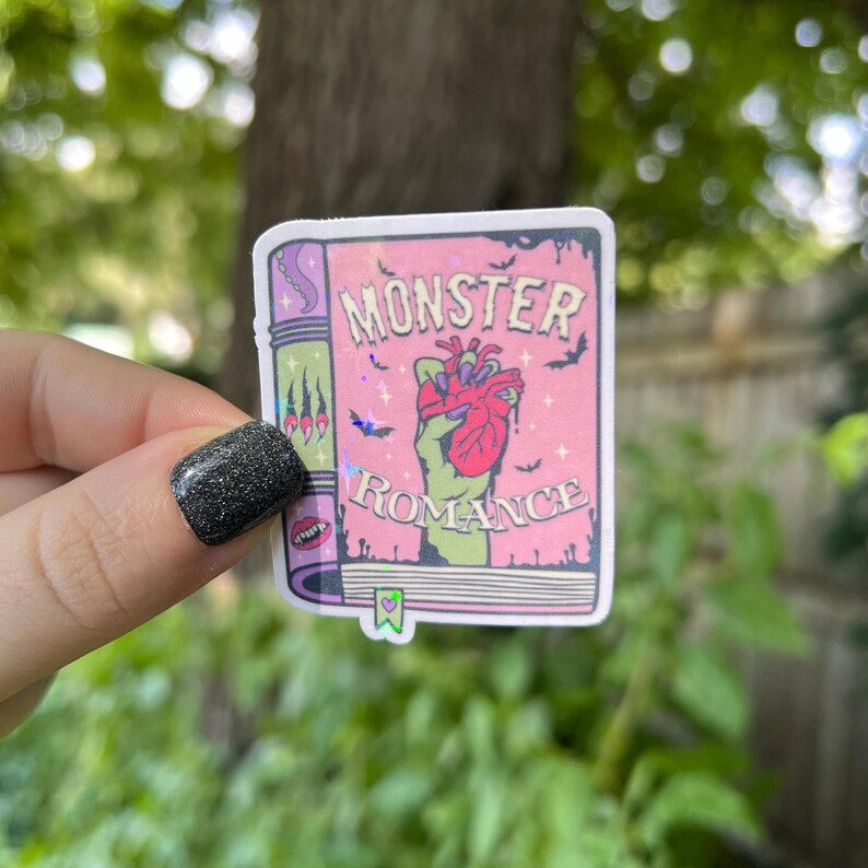 May include: A pink and green sticker with a book cover design. The book cover says "Monster Romance" and features a green hand holding a bloody heart.