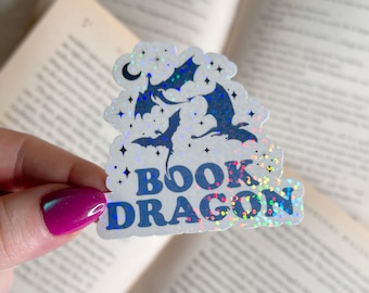 May include: A holographic sticker with a whimsical design featuring three dark blue dragons, stars, and a crescent moon. The words "BOOK DRAGON" are printed in blue. The sticker has a glittery, iridescent finish.