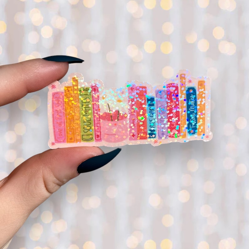 Puede incluir: Una pegatina de una estanter&iacute;a con una variedad de libros coloridos, cada uno con un t&iacute;tulo diferente. Los libros est&aacute;n cubiertos de purpurina iridiscente y tienen un fondo rosa. La pegatina es perfecta para a&ntilde;adir un toque de fantas&iacute;a a cualquier superficie.