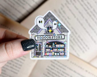 May include: A white sticker with a spooky bookstore illustration. The building is gray with purple accents and a sign that reads "BOOOKSTORE". Two ghosts are floating above the building.