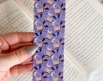 Puede incluir: Marcapáginas morado con un patrón repetido de libros abiertos, murciélagos y lunas crecientes.