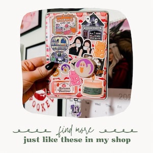May include: A pink and white planner with a variety of stickers, including a ghost, a gummy bear, and a book store. The planner has a calendar with the month of October and the year 2022. The planner has a charm attached to it with the letter M.