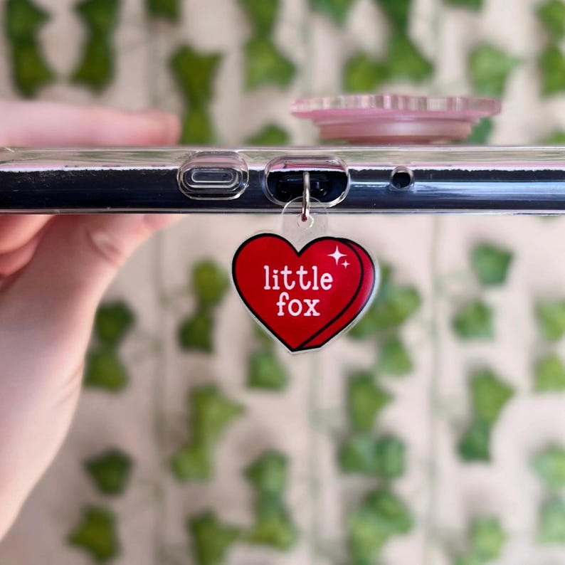 Puede incluir: Un charm de tel&eacute;fono en forma de coraz&oacute;n rojo con el texto "little fox" en letras blancas. El charm est&aacute; unido a una funda de tel&eacute;fono transparente.