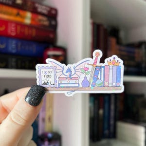 May include: A holographic sticker featuring a miniature bookshelf design. The sticker includes a dragon, sword, crown, and books. The text "I DO NOT YIELD" is displayed on a framed picture. The color palette includes pink, blue, and purple.