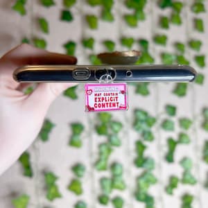 Peut inclure: Un porte-clés de téléphone rose et blanc avec le texte "WARNING MAY CONTAIN EXPLICIT CONTENT" et des roses rouges autour de la bordure.