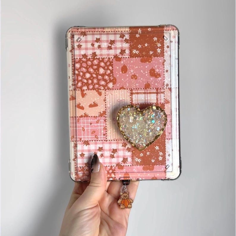 May include: A rectangular tablet case featuring a patchwork design in shades of orange, pink, and white with patterns of leaves, pumpkins, and plaid. A heart-shaped, iridescent, glittery attachment is centred, and a small bear charm hangs below.