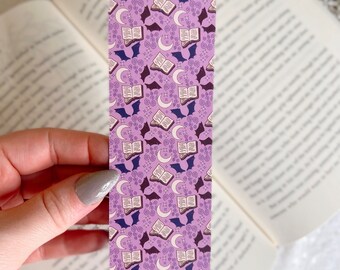 Puede incluir: Marcapáginas morado con un patrón repetido de libros abiertos, lunas crecientes y murciélagos. El patrón está en tonos de morado, marrón y blanco.