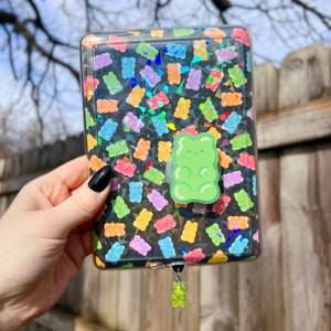May include: A clear plastic case with a gummy bear design. The case has a green gummy bear charm attached to it.