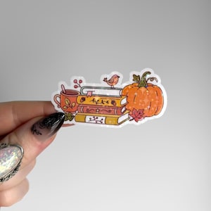 May include: A holographic sticker featuring a stack of three books, a steaming mug, a small bird perched on top, and a large orange pumpkin. The illustration includes autumn-themed details like acorns and leaves with a sparkling star effect.