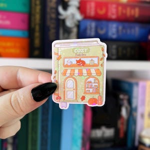 May include: A sticker featuring a cozy bookstore design. The sticker has a light green background with a bookstore facade, an orange awning, and the words "COZY Bookstore" at the top. The sticker includes a fox, pumpkins, and fall leaves.