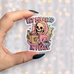 May include: Holographic sticker featuring a skeleton in a pink hoodie reading a book. The sticker has the text "LET ME READ IN PEACE" in a colorful font. A blue mug and a pillow are also depicted.