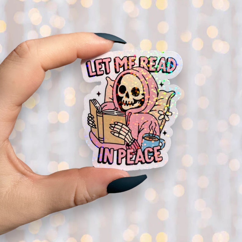 May include: Holographic sticker showing a skeleton in a pink hoodie reading a book. The sticker has the text "LET ME READ IN PEACE" in a colourful font. A blue mug and a pillow are also depicted.