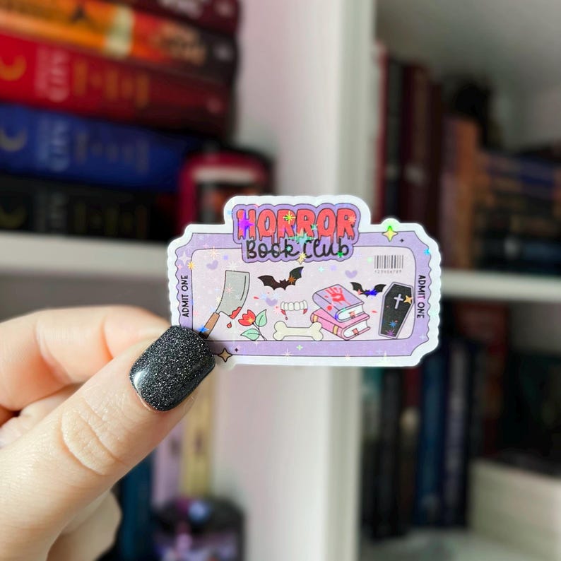 May include: A sticker with a ticket design, featuring the text "HORROR Book Club." The sticker includes illustrations of a cleaver, bats, books, a bone, and a coffin. The background is light purple with a holographic effect.