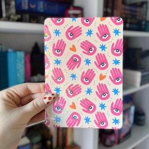 May include: A rectangular notebook with a cream background, patterned with pink hands with eyes, blue stars, and orange hearts. The notebook is held in a person's hand. The design has a mystical theme.