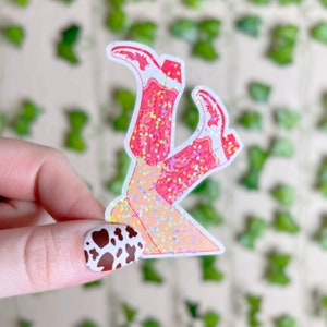 May include: A sticker of a pair of pink cowboy boots with glitter, the boots are crossed at the ankles and the sticker is on a white background.