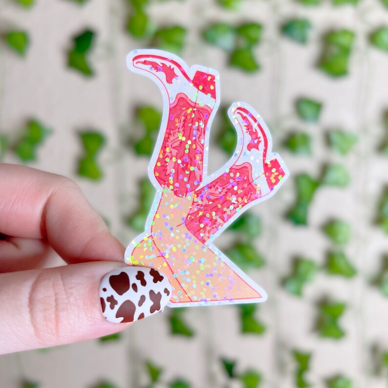 May include: A sticker of a pair of pink cowboy boots with glitter, the boots are crossed at the ankles and the sticker is on a white background.