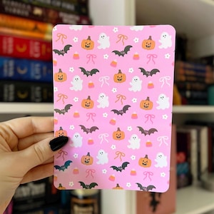 May include: A pink rectangular card with a repeating pattern of Halloween-themed illustrations. The design includes orange pumpkins, white ghosts, black bats, white flowers, pink bows, and candy corn. The card is held by a hand with a black painted nail.
