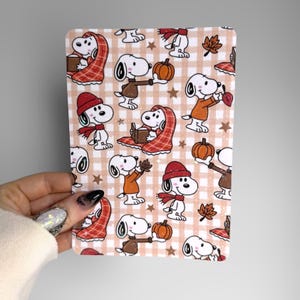 May include: A rectangular sheet featuring a repeating pattern of Snoopy characters on a light beige and white gingham background. The cartoon dog is depicted in various autumn-themed poses, including wearing hats and scarves, holding pumpkins, and reading books.