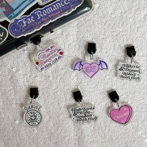 May include: Six different book club charms with purple, pink and black accents. The charms feature text that says "Monster Romance Book Club", "I Like My Men Tall, Dark & Illyrian", "Valkyrie & Dragon Riders Book Club", "Suriel", "High Lords & Wing Leaders Book Club", and "Cassian".