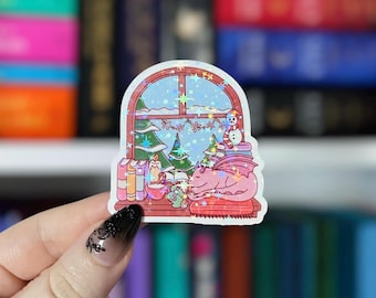 May include: A holographic sticker featuring a winter scene with a window, Christmas trees, and a snowman. A pink dragon is sleeping on a pillow. Books and candles are also visible. The sticker is held by a hand with a black manicure.