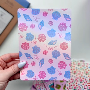 May include: A rectangular card with a gradient background of pink, white, and blue. The card features a pattern of blue and pink seashells and small bows. The card is held by a person.
