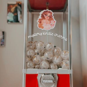 May include: A red and white gumball machine filled with clear plastic spheres containing gold accents. A sticker of a woman with glasses and the text "mystery kindle charms" is attached to the machine.